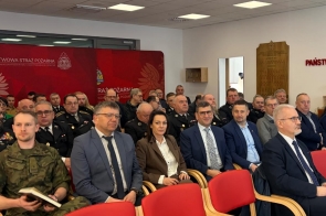 Kilka rzędów osób siedziących  na czerwonych krzesłach, wśród nich strażacy w czarnych mundurach galowych oraz osoby w garniturach i marynarkach. . W tle czerwona ścianka. Na suficie widoczne panele oświetleniowe.