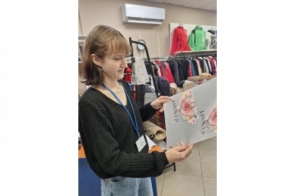 Młoda kobieta stoi w pomieszczeniu przypominającym sklep z odzieżą. Trzyma w dłoniach otwartą kartkę z ilustracją kwiatów i napisem. W tle widoczne są wieszaki z ubraniami, półki oraz klimatyzator na ścianie. Kobieta ma na szyi identyfikator.