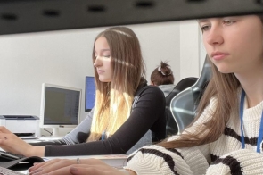 Dwie kobiety siedzą przy biurku i pracują przy laptopach. Na blacie znajdują się dokumenty oraz kubek. W tle widoczne są ściany pomieszczenia i elementy wyposażenia biurowego.