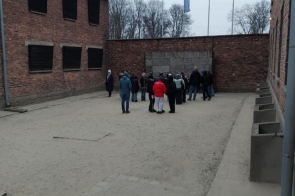 Zamknięty dziedziniec otoczony ceglanymi budynkami z ciemnymi oknami. Na środku placu stoi grupa osób ubranych w zimowe kurtki, zwróconych w stronę ściany z dużymi betonowymi płytami. W tle widoczne są maszty flagowe oraz drzewa bez liści. Po prawej stronie znajdują się betonowe elementy ustawione w rzędzie wzdłuż budynku.