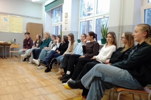 Sala lekcyjna z jasnymi ścianami i dużymi oknami. Kilkanaście osób siedzi w rzędzie na krzesłach ustawionych pod ścianą. Na parapetach stoją rośliny doniczkowe, a na ścianie wiszą plansze z wykresami matematycznymi. Po lewej stronie znajduje się stolik z poczęstunkiem i czajnikiem.