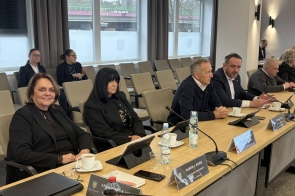 Radni siedzą przy długim stole w sali sesyjnej. Przed nimi znajdują się mikrofony, tabliczki z imionami i nazwiskami, tablety, filiżanki z kawą oraz butelki z wodą. W tle widoczne są rzędy krzeseł oraz duże okna z roletami i zasłonami. Radni mają na sobie stroje formalne.