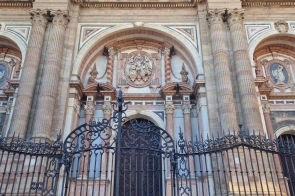 Grupa osób siedzi na schodach przed wysoką, ozdobną bramą z kutego metalu. W tle widać monumentalną fasadę budynku z kolumnami, łukami i bogatymi zdobieniami architektonicznymi. Budowla ma jasną, kamienną elewację oraz symetryczny układ okien i dekoracji.