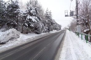 Odcinek drogi przebiegającej przez teren zalesiony w zimowej scenerii. Nawierzchnia jest odśnieżona, po bokach zalega śnieg, a drzewa pokryte są białą warstwą. Po prawej stronie widoczna zielona barierka ochronna oraz słup energetyczny z transformatorem.
