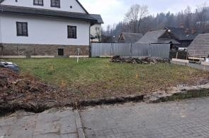 Ogrodzony teren z trawnikiem oraz pasem odsłoniętej ziemi wzdłuż ogrodzenia, przed budynkiem o jasnej elewacji i kamiennej podmurówce. W tle widoczne są inne zabudowania oraz drzewa, a na pierwszym planie fragment chodnika z kostki brukowej.