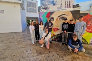 Grupa młodych osób pozuje do zdjęcia na placu wyłożonym kamiennymi płytami. W tle widoczny jest kolorowy mural przedstawiający twarz kobiety z czerwonymi ustami. Część osób stoi, dwie osoby kucają z przodu.