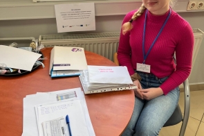 Dziewczyna siedzi przy okrągłym stole, na którym leżą dokumenty, segregator i długopis. Ma na szyi identyfikator na niebieskiej smyczy. Za nią znajduje się okno oraz kartka informacyjna dotycząca stażu uczniowskiego.