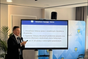 Przy dużym ekranie stoi mężczyzna w garniturze i trzyma mikrofon.  W tle widoczna jest ścianka z herbami Powiatu Cieszyńskiego.