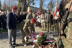 Żołnierz w czerwonym berecie pochyla się nad wieńcem z czerwonych i białych kwiatów. Obok stoi starszy mężczyzna w garniturze i berecie. W tle widać żołnierzy, flagi oraz grupę ludzi, a na pierwszym planie stoi żołnierz z karabinem.