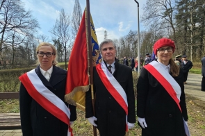 Trzy osoby stoją na zewnątrz przy sztandarze, ubrane w czarne płaszcze z biało-czerwonymi szarfami, podczas uroczystości z udziałem innych uczestników w tle.