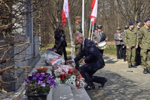 Mieczysław Szczurek Wicestarosta Cieszyński klęka przy pomniku i składa wiązankę kwiatów, a w tle stoją żołnierze, poczet flagowy i orkiestra.