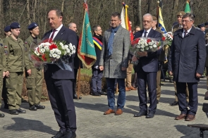Mężczyźni w garniturach trzymają wiązanki kwiatów podczas uroczystości, a za nimi stoją żołnierze i poczty sztandarowe.