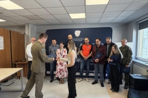 Mężczyzna w mundurze wręcza dokument młodej kobiecie, a za nimi stoi grupa osób trzymających teczki, obserwujących uroczystość.