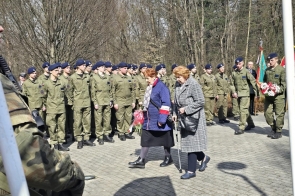 Dwie starsze kobiety idą z wiązanką kwiatów, a za nimi stoi szpaler młodzieży w mundurach podczas uroczystości.
