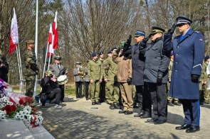 Funkcjonariusze w mundurach stoją na baczność i oddają salut, a obok widać żołnierzy, orkiestrę oraz złożone kwiaty przy pomniku.