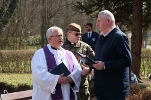 Ksiądz w białej szacie z fioletową stułą rozmawia z mężczyzną trzymającym dokumenty, obok stoi żołnierz w mundurze.