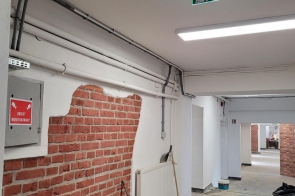 Pomieszczenie w trakcie prac remontowych – białe ściany z miejscami odsłoniętej cegły, instalacje pod sufitem oraz sprzęt i materiały budowlane w środku.