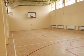 Sala wewnątrz budynku – jasne ściany, drewniana podłoga oraz oznaczenia na podłodze, pod sufitem widoczne instalacje.