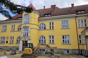 Zabytkowy, żółty budynek szkoły w trakcie remontu. Przed nim stoi koparka i materiały budowlane, widać prowadzone prace.