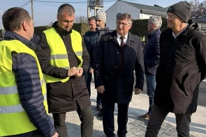 Grupa mężczyzn stoi na nowym chodniku i rozmawia, część z nich ma na sobie kamizelki odblaskowe, a w tle widać zabudowania i elementy infrastruktury.