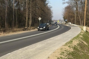 Zakręt asfaltowej drogi z chodnikiem po prawej stronie, przy której jadą samochody, otoczony drzewami i liniami energetycznymi.