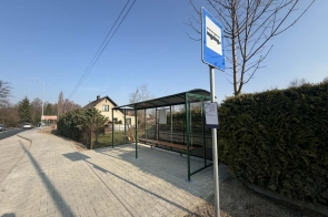 Przystanek autobusowy z zadaszoną wiatą i ławką przy chodniku, obok znak przystanku, a w tle dom i ogrodzenie z żywopłotu.