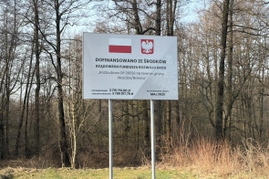 Tablica informacyjna o dofinansowaniu z Rządowego Funduszu Rozwoju Dróg dla rozbudowy drogi w gminie Skoczów/Brenna, ustawiona przy lesie.