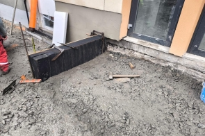 Fragment elewacji budynku z odsłoniętym fundamentem. Przy ścianie czarna izolacja. Na ziemi żwir i nierówne podłoże, widoczny fragment drzwi wejściowych.