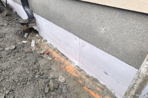 Fragment elewacji budynku z odsłoniętą warstwą izolacji przy fundamencie. Na ścianie szara powierzchnia, poniżej pas ocieplenia. Na ziemi żwir i drobne kamienie.