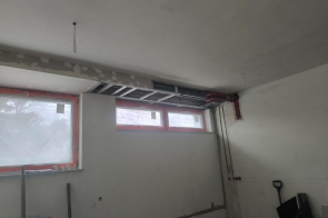 Wnętrze pomieszczenia z oknem pod sufitem. Na ścianie widoczne przewody elektryczne i fragment instalacji. Sufit jasny, ściany w trakcie prac, brak wyposażenia.