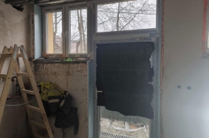 Wnętrze pomieszczenia z otwartymi drzwiami prowadzącymi na zewnątrz. Widoczne okno, ściany i fragment wyposażenia. Na podłodze beton, przy drzwiach próg i żwir na zewnątrz.