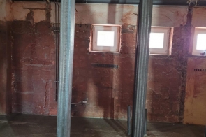 Pomieszczenie w remoncie z surowymi ścianami, metalowymi profilami i rurami przy suficie. W środku widać betonową podłogę i elementy instalacji.