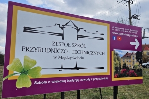 Baner Zespołu Szkół Przyrodniczo-Technicznych w Międzyświeciu ustawiony na zewnątrz. Na fioletowym tle jest nazwa szkoły oraz grafika budynku. Po prawej stronie znajdują się dane adresowe.Obok umieszczono zdjęcie budynku szkoły otoczonego zielenią i kwiatami.