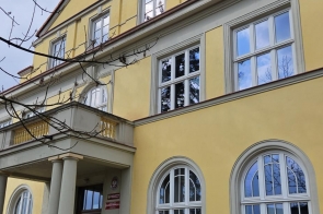 Żółty, kilkupiętrowy budynek z czerwonym dachem i dużymi oknami. Przy wejściu znajdują się schody i kolumny, a wokół rosną drzewa pod pochmurnym niebem.