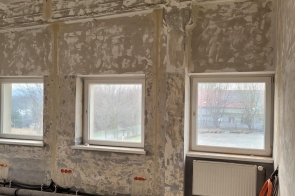 Pomieszczenie  z surowymi, częściowo oczyszczonymi ścianami i widocznymi przewodami przy suficie. Pod oknami biegną rury, a przy jednym z nich znajduje się grzejnik.
