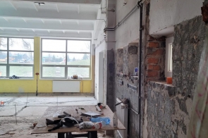 Duże pomieszczenie  z odsłoniętymi ścianami i instalacjami. Na środku stoi stół z narzędziami, a na podłodze leżą materiały budowlane. W tle widać duże okna i grzejnik.