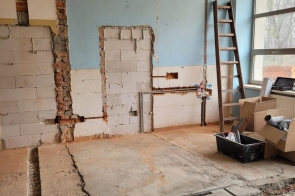 Duże pomieszczenie  z odsłoniętym sufitem i przewodami. Na podłodze leżą płyty i narzędzia, a przy oknach stoją materiały budowlane.