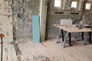 Pomieszczenie z odsłoniętymi ścianami i instalacjami. Na podłodze widać rury i bruzdy, a obok stoi stół z narzędziami i materiałami budowlanymi.