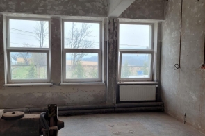 Pomieszczenie  z trzema oknami, surowymi ścianami i widocznymi przewodami. Na podłodze są ślady prac i elementy instalacji, a przy ścianie znajduje się grzejnik