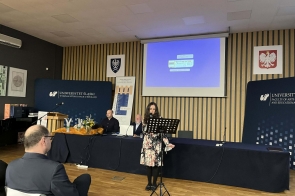Kobieta stoi przy mikrofonie i śpiewa, przed nią ustawiony jest pulpit na nuty. Za nią siedzą trzy osoby przy stole, a na ścianie widać ekran projekcyjny i tablice z logo uczelni.