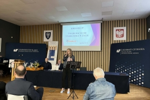 Kobieta śpiewa do mikrofonu przed ekranem z prezentacją, a za nią widać logo Uniwersytetu Śląskiego i siedzących uczestników.