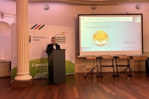 Mężczyzna stoi przy mównicy i przemawia do zgromadzonych. Obok znajduje się ekran z wyświetloną prezentacją „Otwarcie Konferencji”.