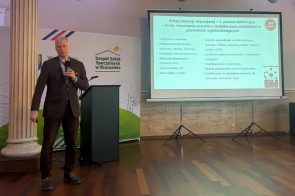 Mężczyzna stoi przy mównicy i przemawia do uczestników, trzymając mikrofon. Obok wyświetlana jest prezentacja.
