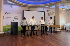 Czterech młodych mężczyzn stoi przy ustawionych instrumentach perkusyjnych i gra na nich, trzymając pałeczki w dłoniach. W tle znajduje się ekran projekcyjny oraz banery instytucji.