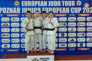 Czterech młodych mężczyzn w strojach judo stoi na podium, prezentując zdobyte medale. Jeden z nich ma złoty medal, pozostali srebrne i brązowe.