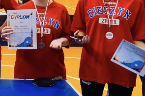 Dwie dziewczyny w czerwonych koszulkach stoją przy stole do tenisa stołowego. Mają na szyjach medale i trzymają dyplomy.