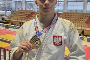 Młody mężczyzna w stroju judo stoi na trybunach hali sportowej i pokazuje medal trzymany w dłoni. W tle widoczna jest mata do walki oraz trybuny.