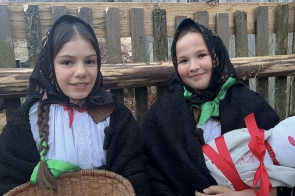 Dwie dziewczynki siedzą obok siebie, ubrane w stroje ludowe z chustami na głowie. Jedna trzyma koszyk, a druga lalkę, a w tle widoczny jest drewniany płot.