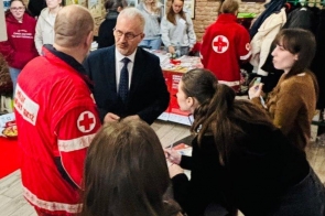 Grupa osób w pomieszczeniu rozmawia przy stoisku informacyjnym. Widoczni są wolontariusze w czerwonych strojach oraz uczestnicy wydarzenia.