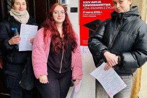 Trzy młode kobiety stoją przy wejściu do budynku, trzymając dokumenty. Obok nich znajduje się czerwony plakat informujący o akcji krwiodawstwa.